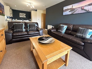A living room with two dark leather sofas, a wooden coffee table, and a kitchen in the background. - 2 bedroom accommodation in Whitby (Whitby)