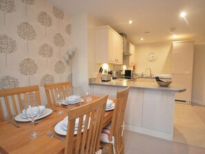 A dining area with a wooden table set for four, a kitchen with modern appliances, and a clock on the wall. - 2 bedroom accommodation in Whitby (Whitby)