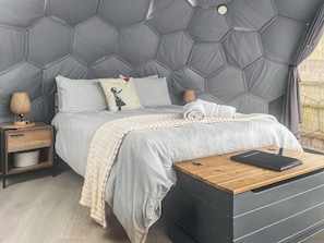 A bedroom with a hexagonal headboard, a bed with white bedding, a bedside lamp, a wooden nightstand, and a black storage unit. - Weets Hill Dome - UKC9598 (Earby)