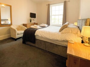 Quarto de hotel com cama, cadeira, mesa de cabeceira, luminária, televisão e janela com cortinas.