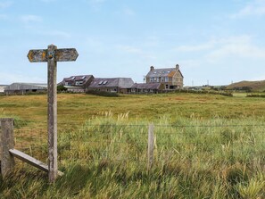A rural landscape with a wooden signpost, a grassy field, and a row of houses with multiple stories and sloped roofs. - 2 bedroom accommodation in Llanengan (Llanengan)
