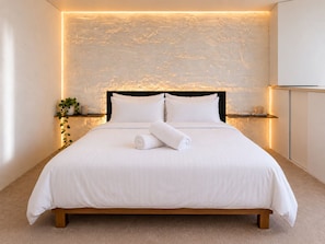 A neatly made bed with white linens and two rolled towels, a bedside table with a plant, and a wall with a textured pattern. - Balcony 1-Bed Apartment Near Sydney Road (Brunswick)