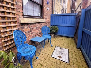 Un petit espace extérieur aménagé avec deux chaises bleues et une table, un treillage en bois et un pot de fleurs.