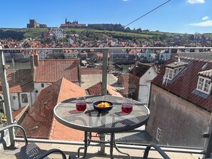 A balcony with a round table, a chair, and a view of a cityscape with buildings and a hilltop structure. - 1 bedroom accommodation in Whitby (Whitby)
