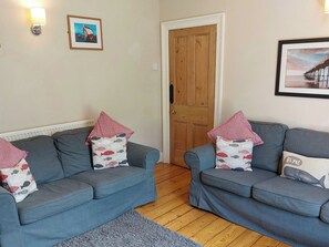 A living room with two grey sofas, each with patterned pillows, a wooden door, and framed pictures on the walls. - 4 bedroom accommodation in Whitby (Whitby)