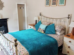 Een bed met een blauwgroene spreidek, kussens met een bloemmotief en een wit metalen hoofdeinde. Er staan nachtkastjes met lampen en een klein lade. In de hoek is een open haard zichtbaar.