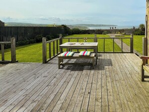 Een houten terras met een tafel en een bankje, met uitzicht op een gazon en een verre horizon van de zee.