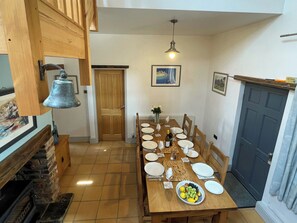 Een eetruimte met een houten tafel gedekt voor zes personen, omringd door stoelen, met een open haard en een klok aan de linkerkant.