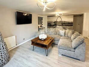 A living room with a grey sofa, a wooden coffee table with a white flower arrangement, and a flat-screen TV mounted on the wall. - 2 bedroom accommodation in Whitby (Whitby)