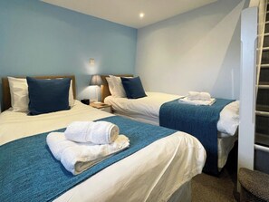 A hotel room with two beds, each with a blue blanket and white linens, a nightstand with a lamp, and a wall-mounted light fixture. - 2 bedroom accommodation in Whitby (Whitby)