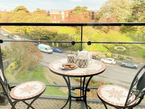 Un balcon avec une table dressée pour le thé, donnant sur une route où circulent des véhicules, et avec un bâtiment visible au loin.