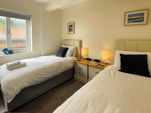 A hotel room with two beds, a bedside table with lamps, a small bedside table with a phone and a remote, and a window with a view of trees. - 3 bedroom accommodation in Whitby (Whitby)