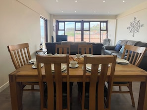 A dining area with a wooden table and chairs, a view of the city through large windows, and a compass wall decoration. - 3 bedroom accommodation in Whitby (Whitby)