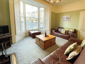 A living room with a brown leather sofa, a wooden coffee table with a bottle and glass, a television on a stand, and large windows with a view of the street. - 3 bedroom accommodation in Whitby (Whitby)