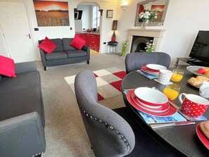 A modern living room with a grey sofa, red cushions, a dining table set with red and white plates, glasses of orange juice, and a fireplace. - 1 bedroom accommodation in Whitby (Whitby)