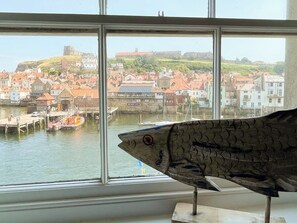 Une sculpture de poisson est placée devant une fenêtre donnant sur un port, avec des bâtiments et une colline en arrière-plan.