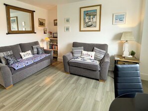Un salon avec deux canapés gris, une table basse en bois et un fauteuil bleu. Les murs sont ornés de tableaux encadrés et une lampe est posée sur une table d’appoint.