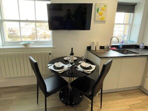 A dining area with a table set for two, a flat-screen TV mounted above, and a window with a view of buildings. - 1 bedroom accommodation in Whitby (Whitby)