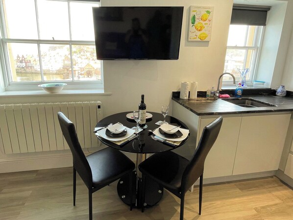 Un coin repas avec une table dressée pour deux personnes, un téléviseur à écran plat fixé au mur au-dessus, et une fenêtre donnant sur des immeubles.