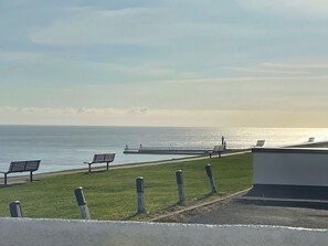 Une vue sur la côte, avec une jetée qui s’avance dans la mer, une zone herbeuse avec des bancs et un bâtiment au premier plan.