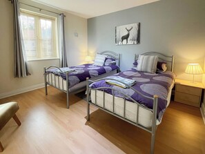 Un dormitorio con dos camas literas de metal, una mesita de noche con lámpara, una ventana con cortinas y un cuadro de un ciervo colgado en la pared.
