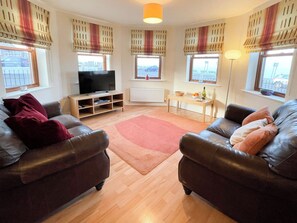A living room with two leather sofas, a television, a small table with a bowl and glasses, and windows with striped curtains. - 2 bedroom accommodation in Whitby (Whitby)