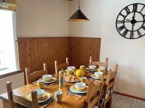 Een eethoek met een houten tafel gedekt voor vier personen, een zwarte wandklok en een raam met zonwering.