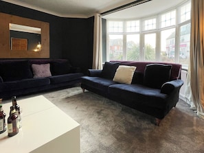 Ett vardagsrum med en soffa i mörk färg, ett vitt soffbord och ett stort fönster med gardiner.
