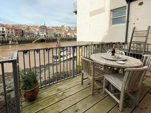 Um deck de madeira com uma mesa e cadeiras, dando vista para um rio com barcos e a paisagem de uma cidade.