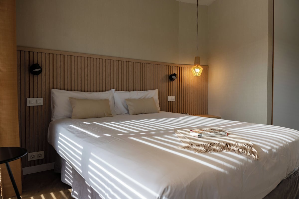 A neatly made bed with a white comforter and several pillows, a bedside table with a lamp, and a wall-mounted light fixture.