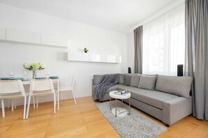 Ein modernes Wohnzimmer mit einem grauen Sofa, einem kleinen weißen Couchtisch und einem Essbereich mit weißen Stühlen und einem Tisch für zwei Personen.
