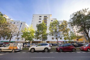 Ein Parkplatz mit mehreren Autos, ein Gebäude mit Balkonen und Bäume im Vordergrund.