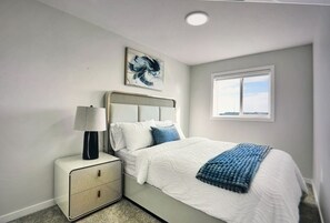 Un dormitorio con cama, mesita de noche, lámpara y una obra de arte en la pared.