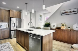 A modern kitchen with wooden cabinets, stainless steel appliances, a central island, pendant lights, and a view of the living area. - Spacious and comfort| Near Airport| Easy Access to Stoney Trail & the Rockies. (calgary)