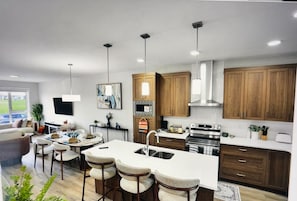A modern kitchen with a central island, bar stools, wooden cabinets, and a large window. - Spacious and comfort| Near Airport| Easy Access to Stoney Trail & the Rockies. (calgary)