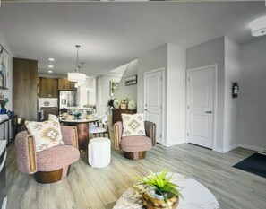 A modern living room with two armchairs, a coffee table, and a dining area with a kitchen in the background. - Spacious and comfort| Near Airport| Easy Access to Stoney Trail & the Rockies. (calgary)