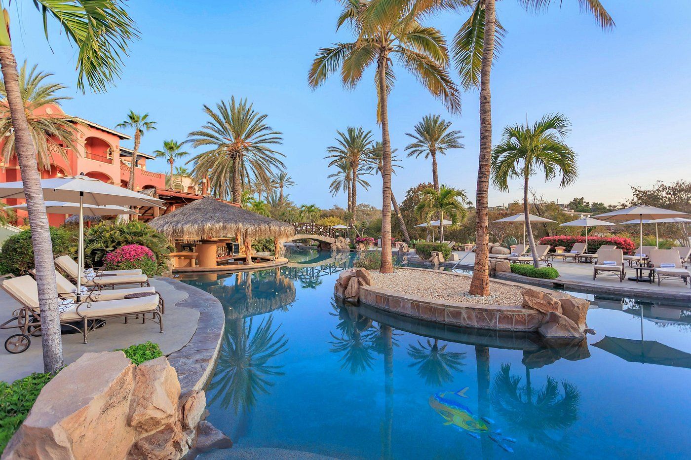 A resort pool area with lounge chairs, palm trees, and a thatched-roof structure.