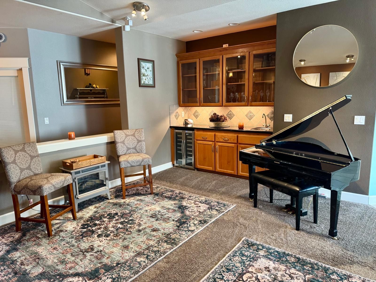 A living room with a grand piano, two armchairs, a fireplace, and a kitchen area with wooden cabinets.