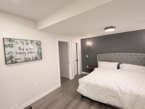 A bedroom with a large bed, a door leading to another room, and a wall-mounted light fixture. - Nolan Hill Garden Retreat walkout basement (Calgary)