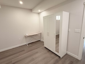 Una habitación con piso de madera, paredes blancas, un armario blanco con puertas y una plancha pequeña apoyada contra la pared.