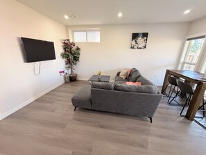 A modern living room with a grey sofa, a flat-screen TV mounted on the wall, a dining table with chairs, and a potted plant. - Nolan Hill Garden Retreat walkout basement (Calgary)