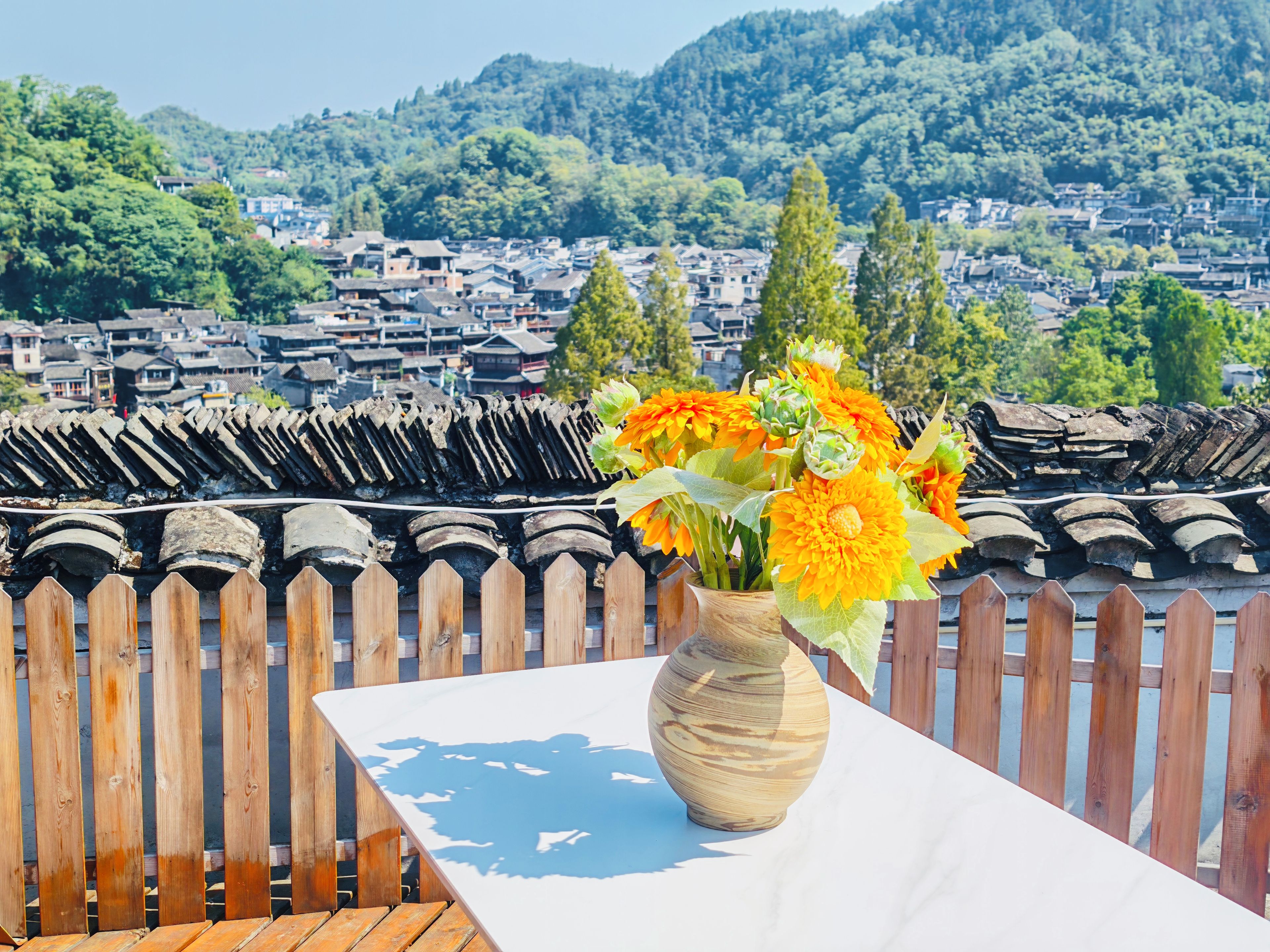 Une clôture en bois surmontée d’un toit traditionnel en tuiles, une table blanche ornée d’un vase de fleurs et une vue sur une ville avec ses maisons et ses arbres.