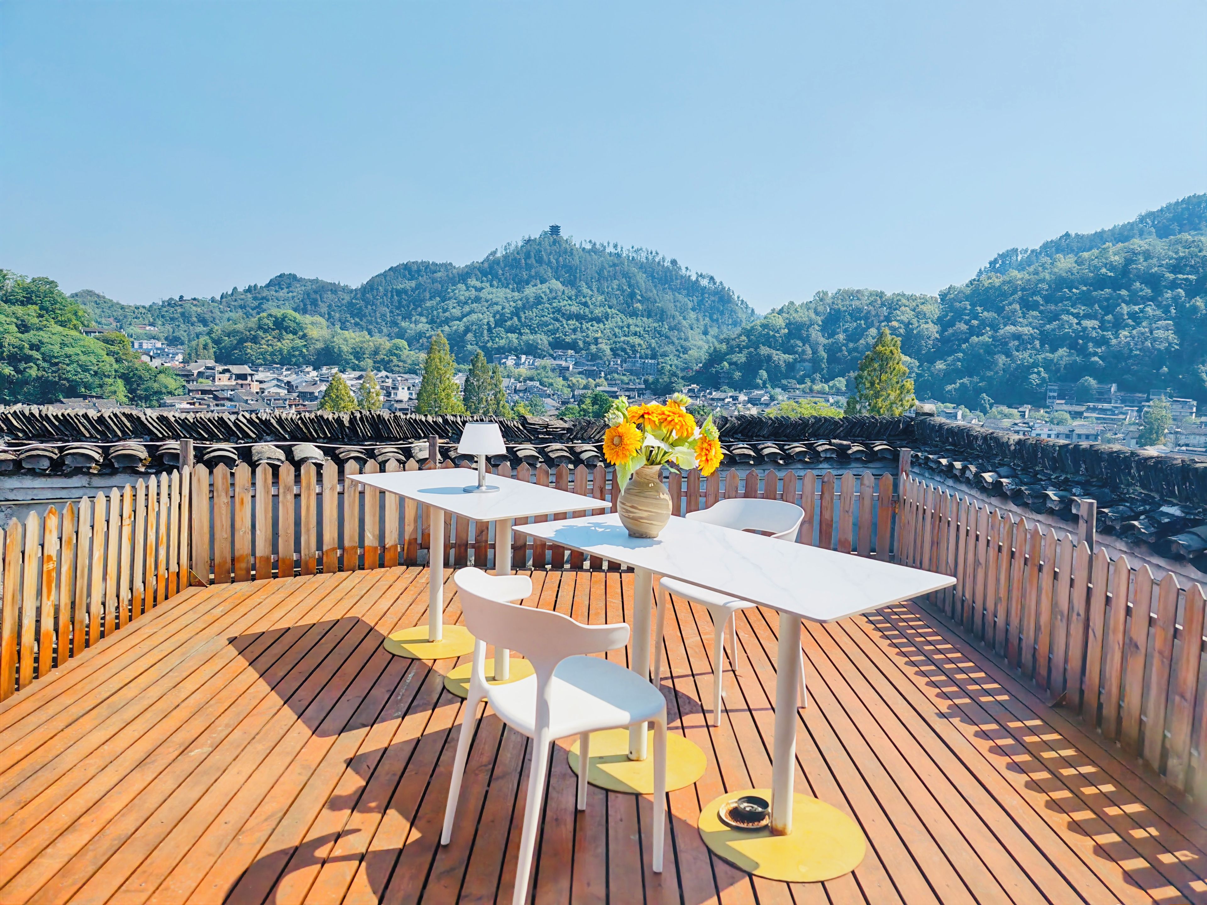 Une terrasse en bois avec une table et des chaises, un vase rempli de fleurs et, en arrière-plan, une vue sur une ville et des collines.