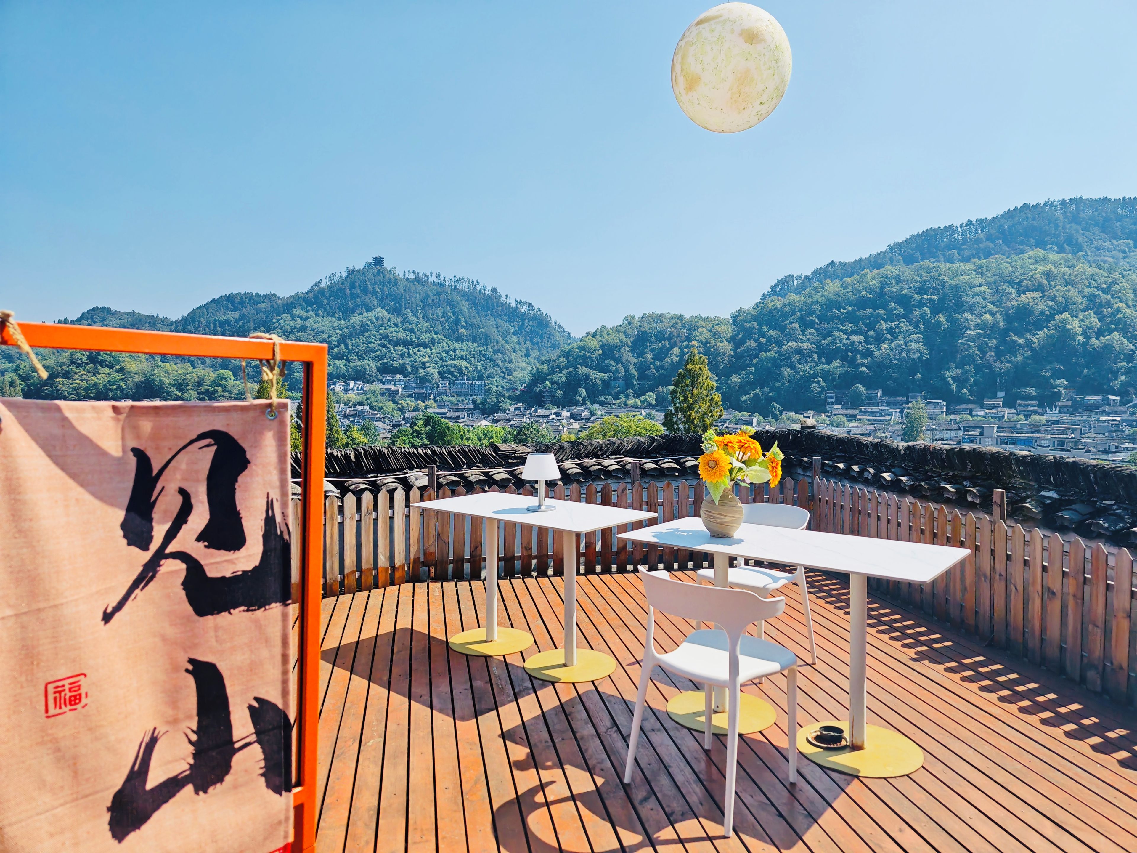 Une terrasse sur le toit avec une plateforme en bois, des tables et des chaises blanches, un vase rempli de fleurs et une vue sur la ville et les montagnes.