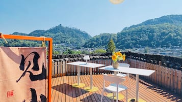 Une terrasse sur le toit avec une plateforme en bois, des tables et des chaises blanches, un vase rempli de fleurs et une vue sur la ville et les montagnes.