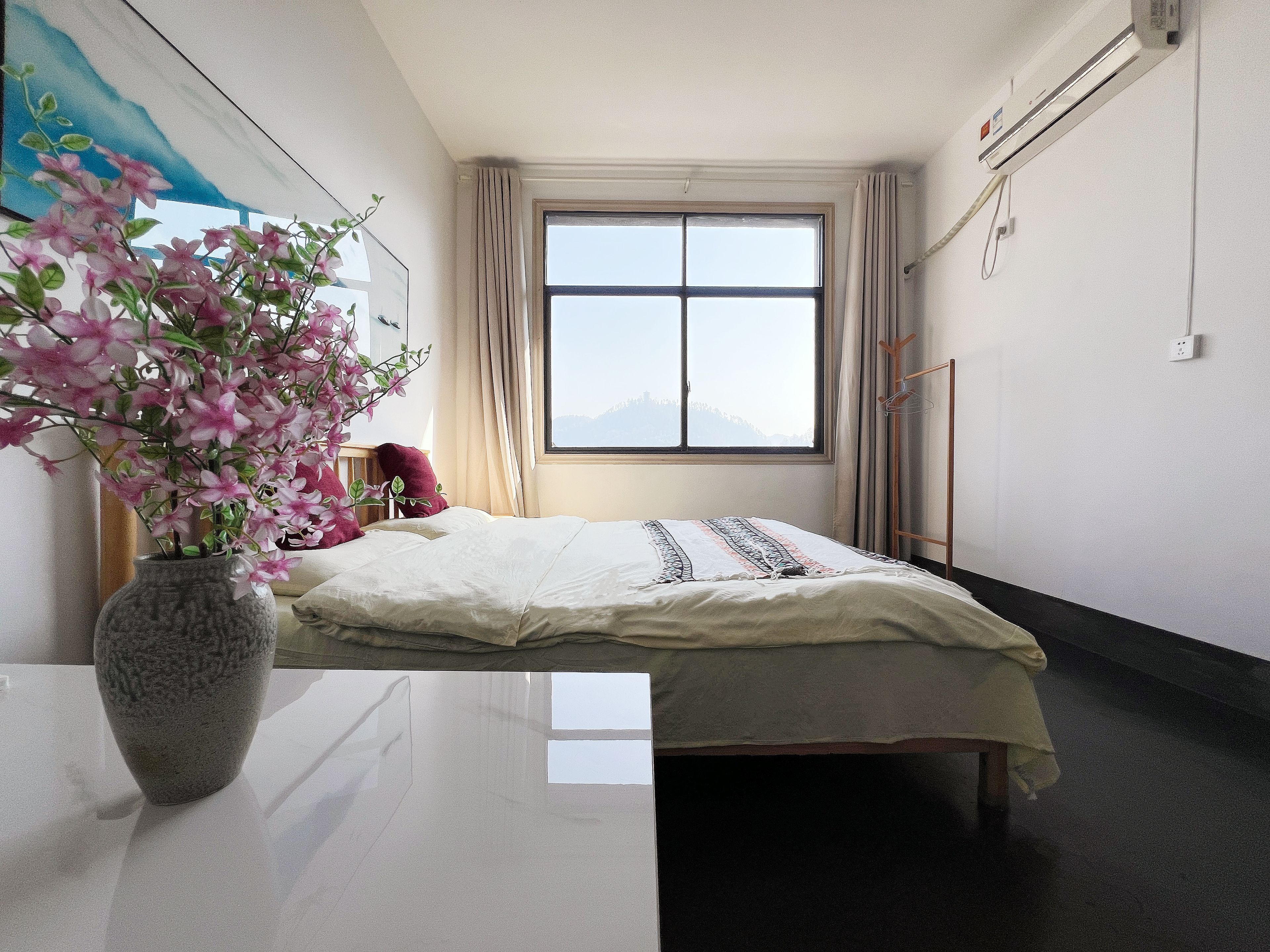 Una habitación de hotel con una cama, un florero con flores, una ventana con cortinas y un aire acondicionado de pared.