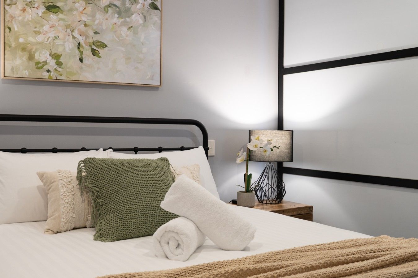 Ein ordentlich bezogenes Bett mit Kissen und einem zusammengerollten Handtuch, eine Nachttischlampe mit einer Orchidee und ein Gemälde an der Wand.