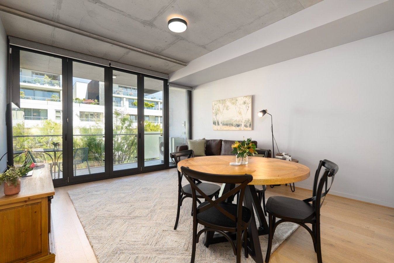 Un intérieur moderne avec une table ronde en bois, des chaises noires, un canapé et une grande fenêtre donnant sur des bâtiments et de la verdure.