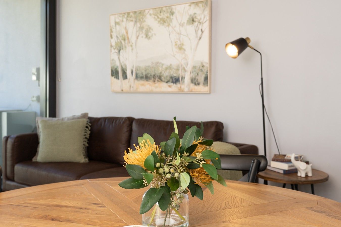 Un salon avec un canapé en cuir marron, une table basse ronde en bois sur laquelle repose un vase en verre transparent contenant des feuilles vertes et des fleurs jaunes, un lampadaire et un tableau accroché au mur.
