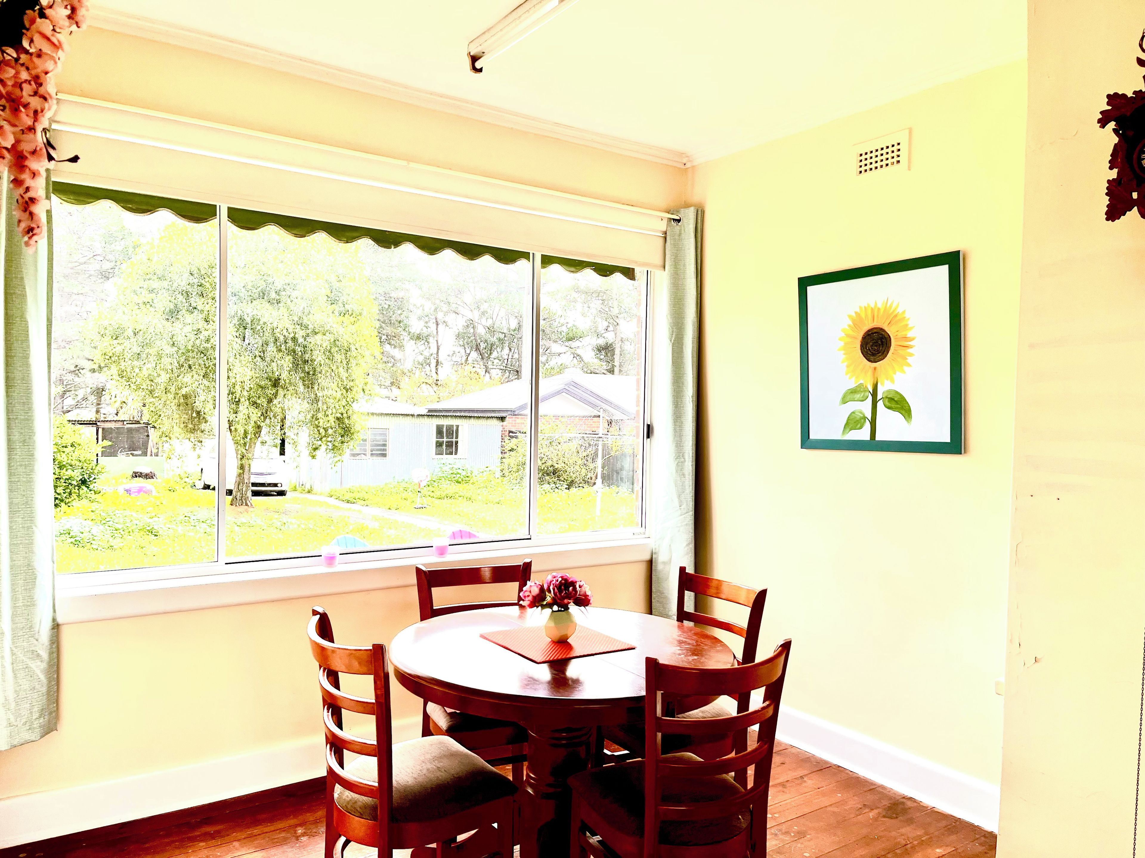 Un coin repas avec une table ronde et des chaises, une fenêtre donnant sur une maison et de la verdure, et un tableau représentant un tournesol accroché au mur.
